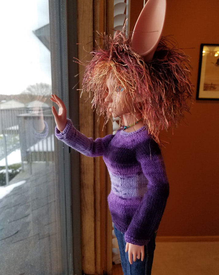 A fennec eared human doll in terracotta colored resin, Featherfall Iruhi, standing looking out the window. She is lit with natural light and is wearing a sweater striped in various shades of purple. The sleeve facing us is mostly dark purple with a band of medium purple around the upper arm from below the shoulder to the elbow.